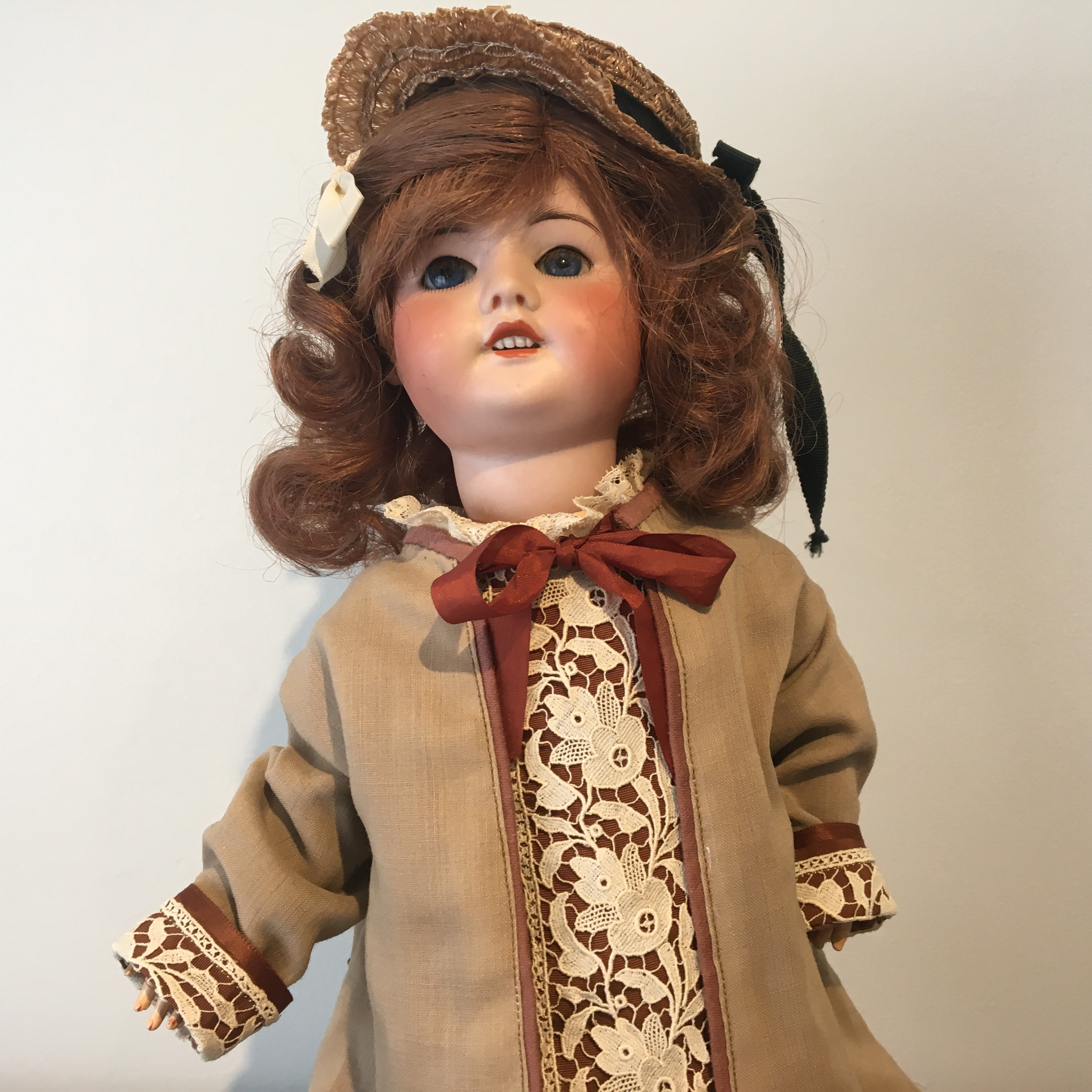 Smiling antique doll with medium brown hair and sleep eyes, with a brown lace-trimmed coat, white skirt, and straw hat