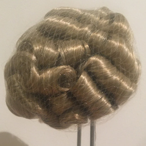 Light brown wig with short, tight curls, contained in a hair net, with a few curls protruding