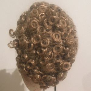 Light brown wig with short, tight curls, contained in a hair net