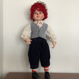 Reimagined Raggedy Andy doll with porcelain child's face painted to resemble the doll and curly yarn hair, with shit shirt, striped white and navy waistcoat, and navy trousers