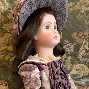 Light-skinned doll in a beige dress with purple trim and elaborate purple ruching, wearing a matching hat over long, straight, dark brown hair
