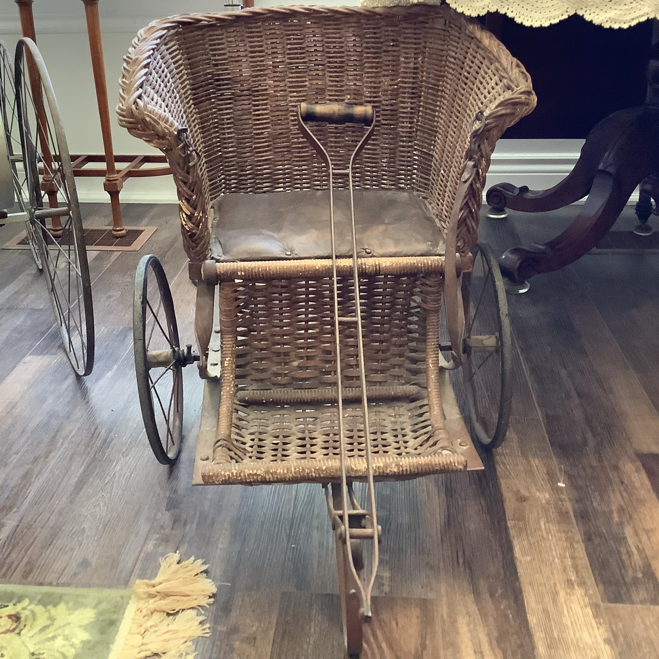 Brown wicker baby carriage with metal wheels, pull handle in front and broken leather seatbelt